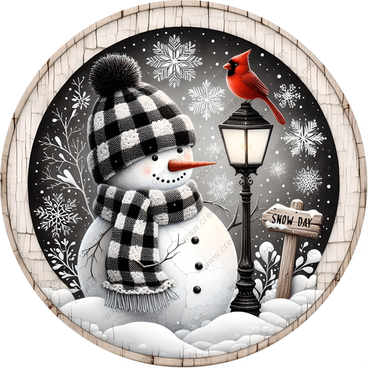 A snowman wearing a black and white checkered hat and scarf stands next to a black lamppost with a red cardinal perched on top. The snowman is surrounded by snowflakes and a snow-covered landscape, with a "Snow Day" sign visible in the background.