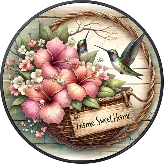 A wicker basket filled with pink and white flowers, with two hummingbirds perched on the basket. The basket is placed on a wooden surface, and there is a sign that reads "Home Sweet Home".