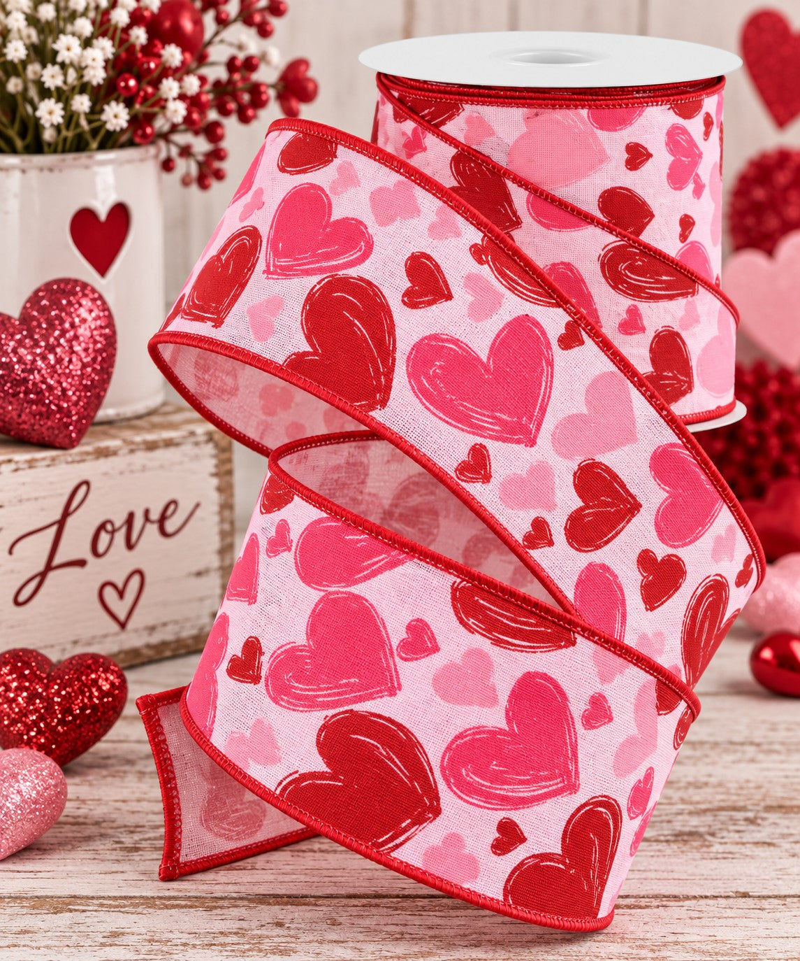 a pink and red heart-patterned ribbon, a white vase with red flowers, and some red and pink glitter hearts, all arranged on a wooden surface.