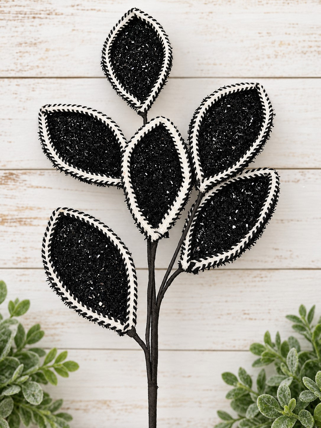 A black and white decorative leaf-shaped ornament with a textured surface, placed on a white wooden surface with green plants in the background.