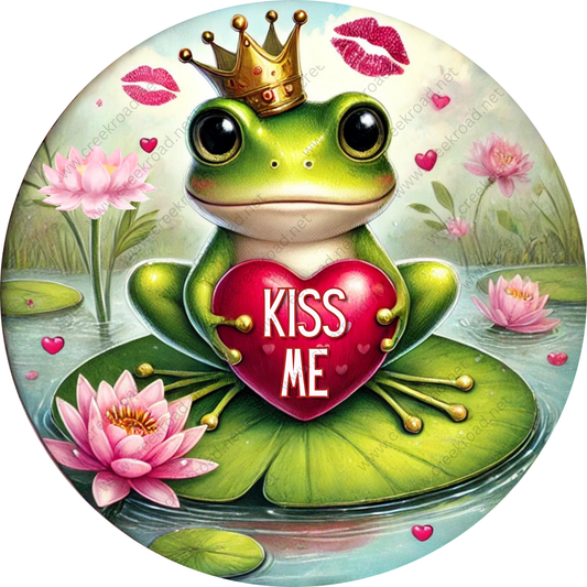 A green frog wearing a crown and holding a heart with the words "KISS ME" is surrounded by pink flowers and lily pads in a pond setting.